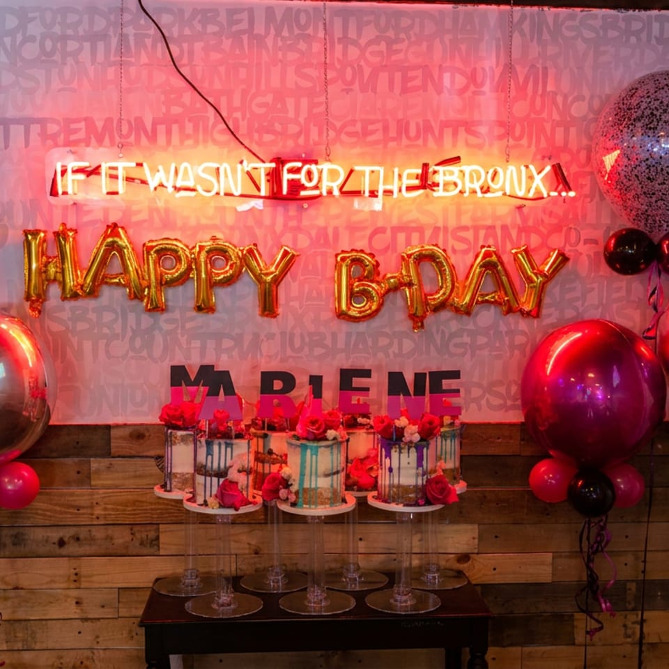 Birthday display with red and black balloons, illuminated “Happy B-Day” sign, and three decorated cakes on a table.