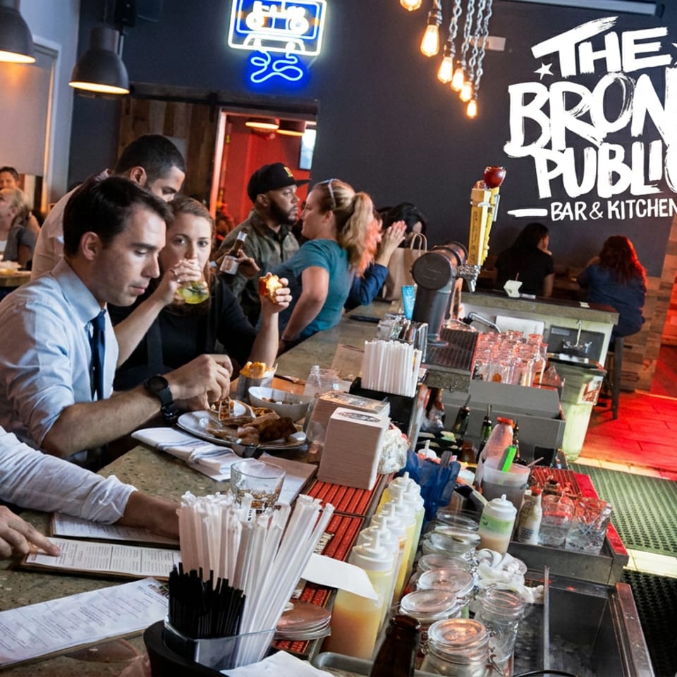 Busy Bronx Public Bar & Kitchen dining area with patrons seated at the bar, enjoying food and drinks under warm pendant lights.