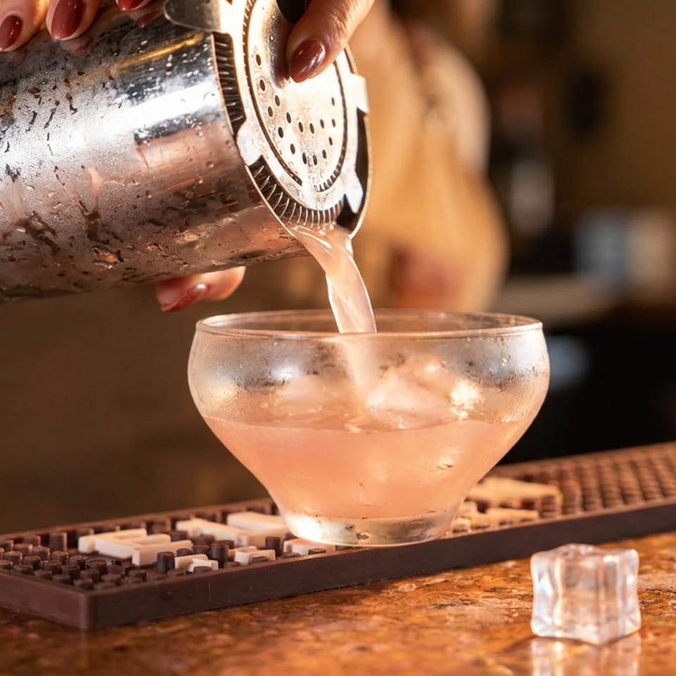 Bartender pours a pale pink cocktail from a metal shaker into an ice-filled coupe glass at a warmly lit bar.