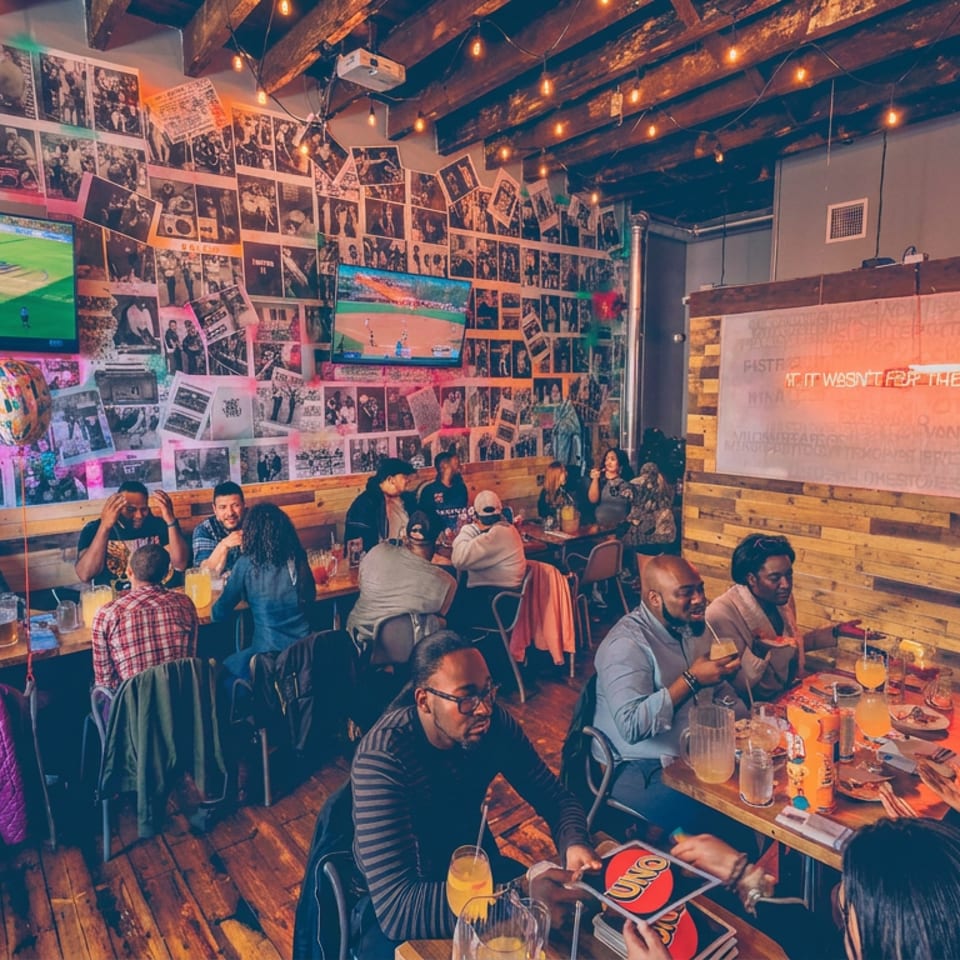 Lively restaurant dining room with groups of diners, colorful drinks, televisions showing sports, string lights, and collage-covered walls.