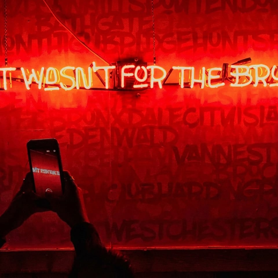 Red neon sign reading “If it wasn’t for the Bronx...” above a person photographing it with a smartphone.