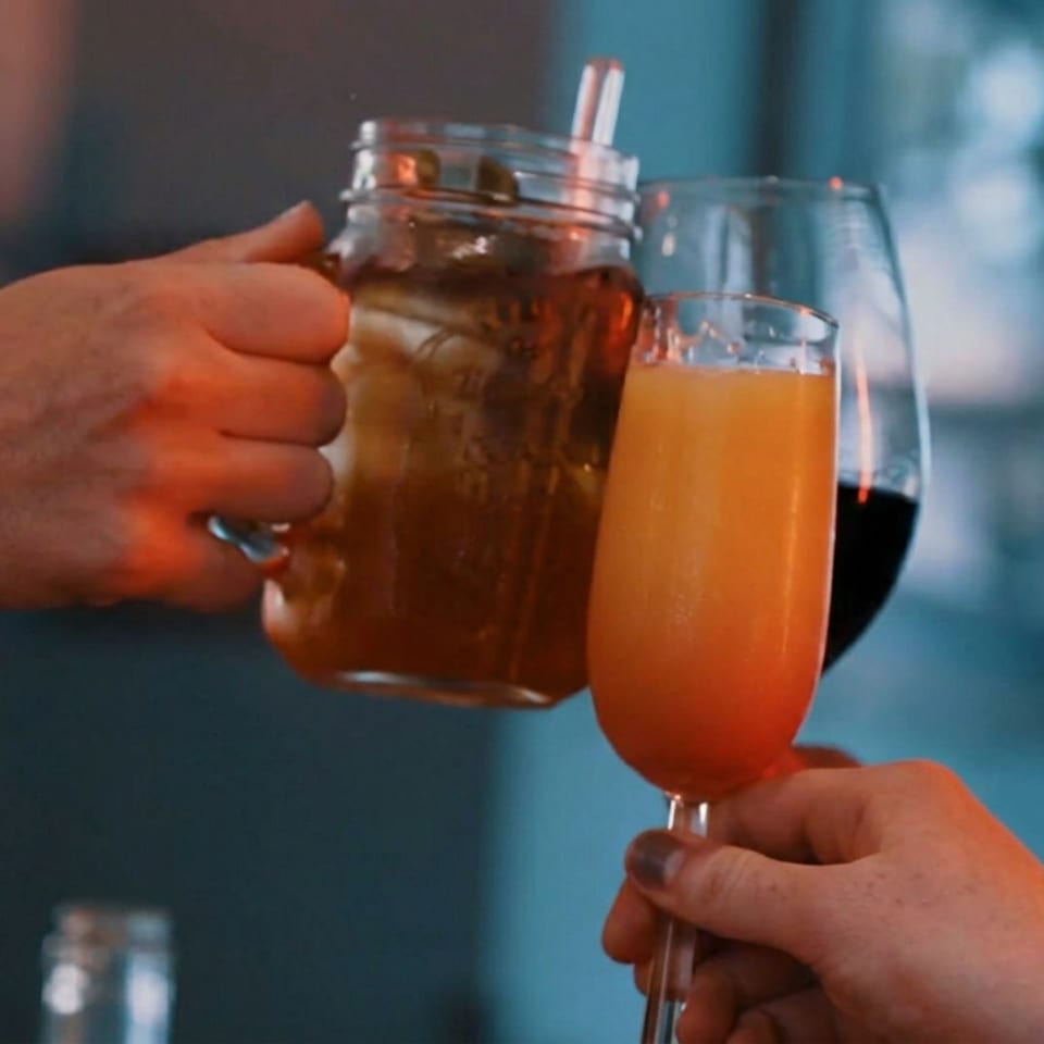 Hands raise a mason jar cocktail and orange drink in a toast at a warmly lit restaurant bar.