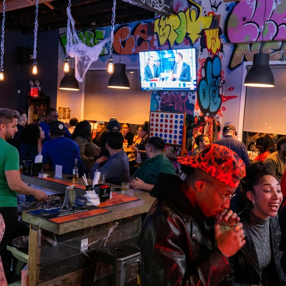 Lively, crowded restaurant bar with diners, hanging lights, graffiti-covered walls, television, and colorful drinks.