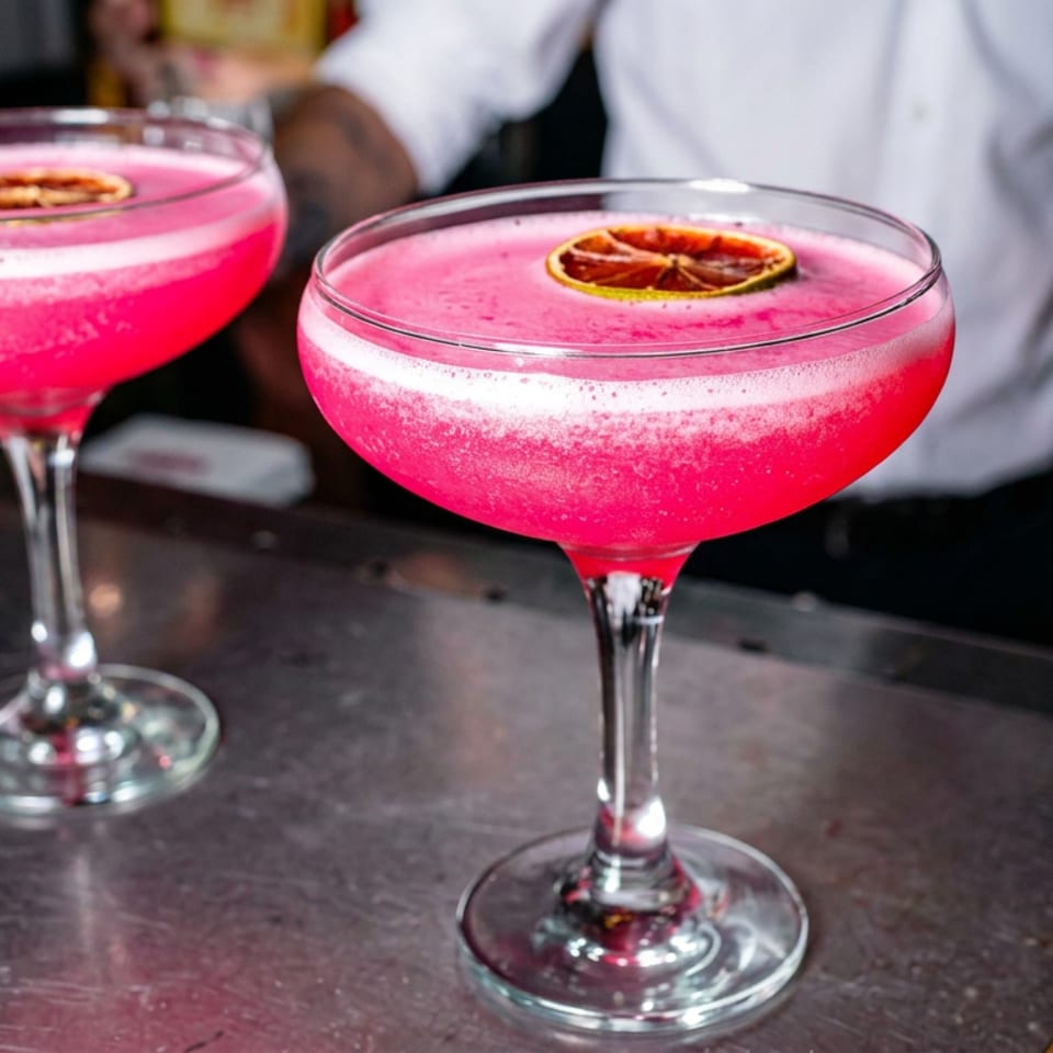 Two bright pink cocktails garnished with dried citrus slices sit on coupe glasses at a restaurant bar.