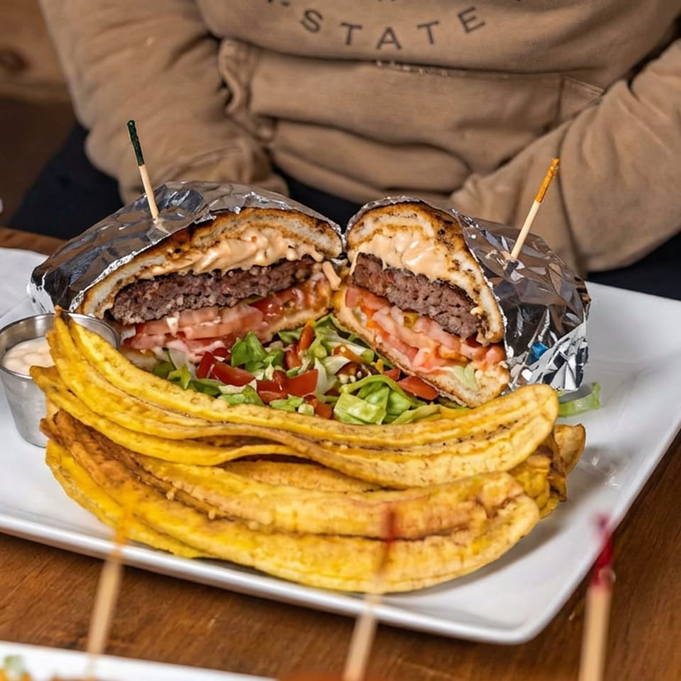 Cut-open burger with beef, cheese, tomatoes, lettuce, and sauce served with golden plantain chips on a white plate.
